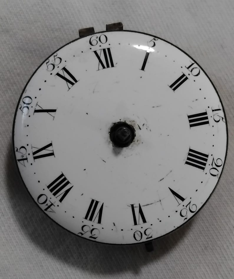 May include: A white clock face with black Roman numerals and minute markers. The clock face has a black outer ring and a central black mechanism. The numerals are arranged around the perimeter of the face.