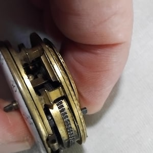 May include: Close-up of a vintage pocket watch mechanism. The watch features a gold-toned metal casing with intricate internal gears and a white enamel face. The mechanism is partially exposed, showcasing the detailed craftsmanship of the antique timepiece.