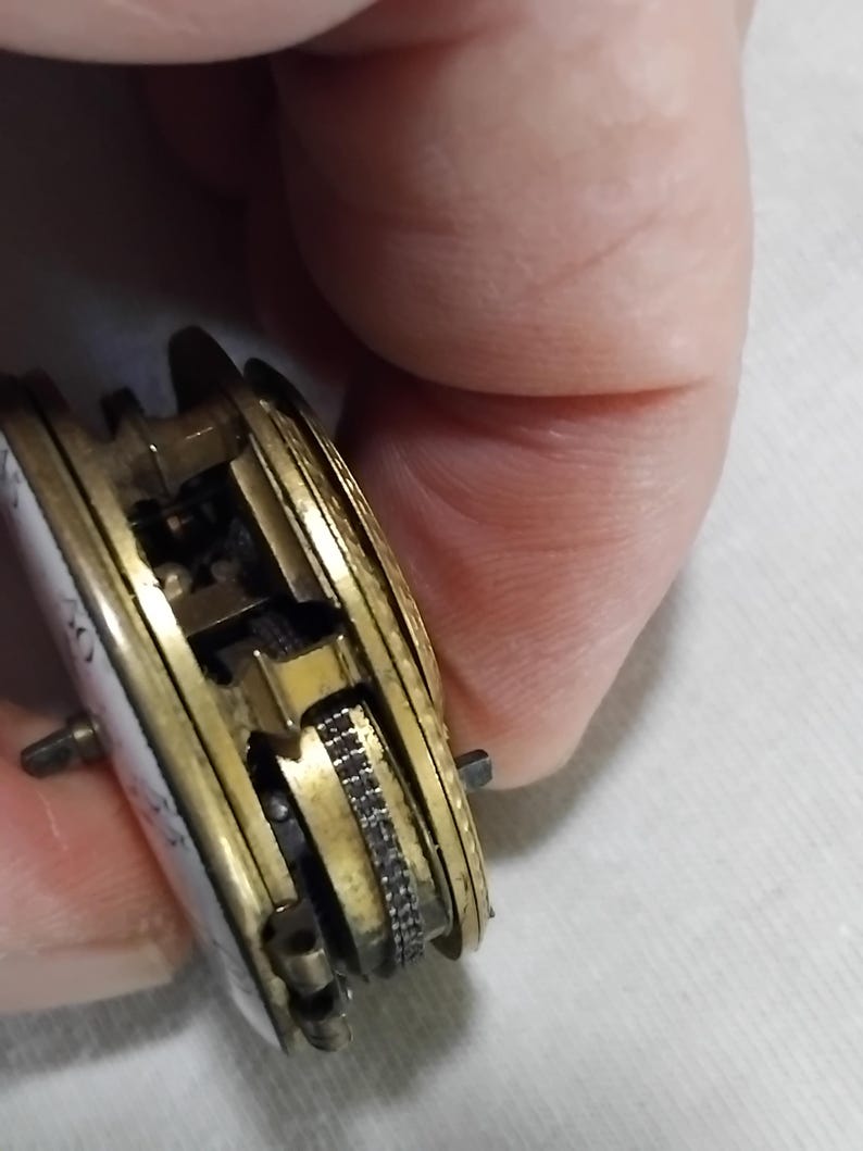 May include: Close-up of a vintage pocket watch mechanism. The watch features a gold-toned metal casing with intricate internal gears and a white enamel face. The mechanism is partially exposed, showcasing the detailed craftsmanship of the antique timepiece.