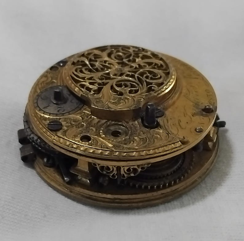 May include: A detailed view of a vintage watch movement. The gold-toned mechanism features intricate engravings and a filigree cover. Visible are gears, screws, and other components, showcasing the craftsmanship of the timepiece. The watch is open, revealing its internal workings.