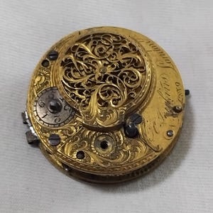 May include: Close-up of a vintage gold pocket watch movement. The watch mechanism features intricate filigree detailing and visible gears. The dial includes Roman numerals and the inscription "London". The watch is a beautiful example of antique craftsmanship.