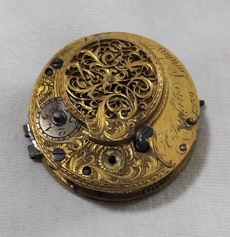 May include: Close-up of a vintage gold pocket watch movement. The watch mechanism features intricate filigree detailing and visible gears. The dial includes Roman numerals and the inscription "London". The watch is a beautiful example of antique craftsmanship.