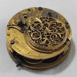May include: Close-up of a vintage gold pocket watch movement. The intricate mechanism features engraved details, gears, and the inscription "T. Watkins London" with the number "2859". The watch has a decorative, openwork design.