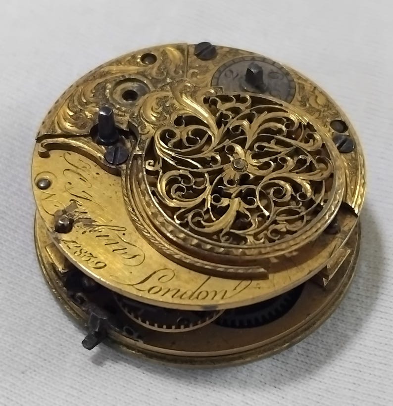 May include: Close-up of a vintage gold pocket watch movement. The intricate mechanism features engraved details, gears, and the inscription "T. Watkins London" with the number "2859". The watch has a decorative, openwork design.