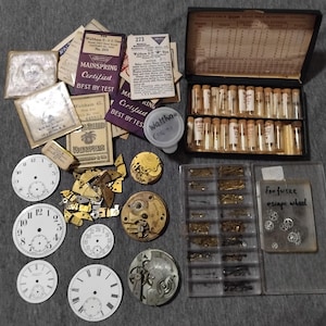 May include: A collection of vintage watch parts, including watch faces, gears, and mainsprings. There are several small glass vials, a black box, and various paper documents. The watch faces are white with black numerals.