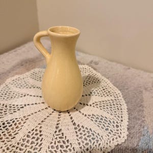 May include: A small, pale yellow ceramic pitcher with a smooth, glossy finish. The vessel features a rounded body, a narrow neck that flares slightly at the top, and a single curved handle attached to the side. The pitcher has a solid, uniform color.