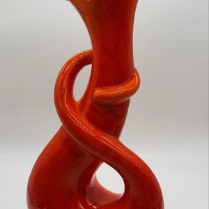 May include: A bright orange ceramic vase with a tall, flared opening and a sculptural, swirling form that wraps around the central neck, creating an open loop design. The surface has a smooth, glossy finish with subtle variations in color intensity.