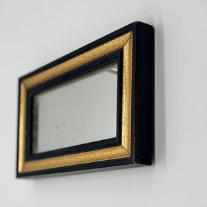 May include: A rectangular mirror featuring a black frame with an inner gold-colored border that has a textured, crackled finish. The mirror is positioned at an angle, displaying a reflective glass surface surrounded by the dual-toned frame.