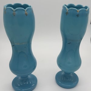 May include: A pair of matching turquoise glass vases with a pedestal base and a scalloped rim. Each vase features a slender, curved body and is decorated with small, gold-colored leaf-shaped accents along the top edge and faint gold detailing.