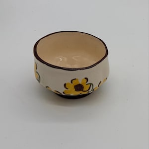 May include: A small, round ceramic bowl with an off-white interior and exterior. The exterior features a hand-painted pattern of yellow flowers with dark brown centers and green stems. The rim and the base of the bowl are trimmed in dark brown.