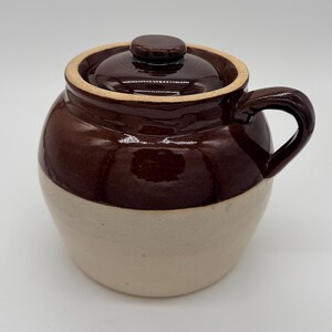 May include: A two-toned ceramic crock with a glossy dark brown glazed upper section and a matte cream-colored lower base. The vessel features a matching dark brown lid with a central knob handle and a single curved handle on the side.