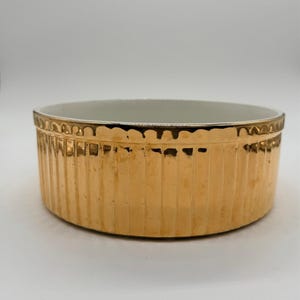 May include: A round ceramic bowl with a metallic gold exterior finish and a white interior. The exterior features a vertical ribbed texture along the sides and a scalloped edge detail around the top rim. The bowl has a wide, shallow profile.
