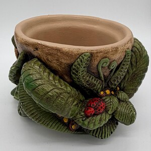 May include: A round, tan-colored ceramic planter featuring a textured exterior decorated with sculpted green leaves, small yellow accents, and a single red ladybug with black spots resting on the foliage. The interior is smooth and light brown.