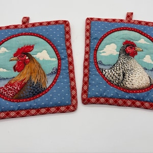 May include: Two square fabric pot holders featuring a blue background with a white pattern. Each has a central circular panel depicting a farm bird against a sky with clouds, framed by a red border with a white cross-hatch pattern and a hanging loop.