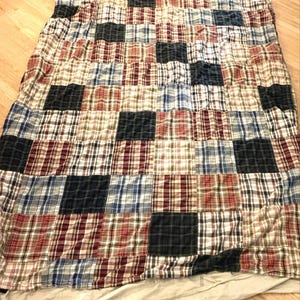 May include: A rectangular patchwork quilt featuring a grid of alternating fabric squares. The design consists of solid dark navy blue squares interspersed with various plaid patterns in shades of red, white, blue, and tan, creating a multi-colored look.