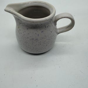 May include: A small ceramic creamer pitcher featuring a light gray base with a speckled pattern of dark blue dots. The pitcher has a rounded body, a curved handle on the side, and a distinct pouring spout at the top rim for liquid dispensing.