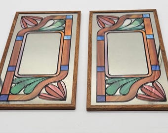 May include: A pair of rectangular wall mirrors framed in wood, each featuring a decorative stained glass border with pink, green, and blue glass segments arranged in a symmetrical pattern surrounding a central reflective glass panel.