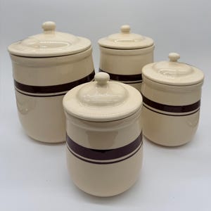 May include: A set of four cream-colored ceramic canisters with matching lids, each featuring a single dark brown horizontal stripe around the center. The canisters vary in size, with one large, one medium, and two smaller containers arranged together.