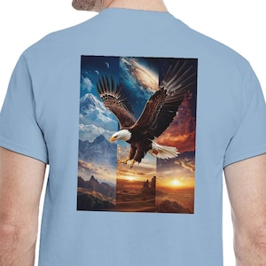 Könnte beinhalten: Hellblaues T-Shirt mit einer detaillierten Grafik eines Weißkopfseeadlers im Flug vor einer Berg- und Sonnenuntergangskulisse. Der Adler hat braune und weiße Federn. Das T-Shirt ist aus weichem Material.