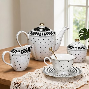 Puede incluir: Un juego de té de porcelana blanca con un patrón de lunares negros y detalles de rayas negras. El conjunto incluye una tetera, una lechera, un azucarero con tapa y una taza de té sobre un platillo. Cada pieza tiene un pequeño pomo dorado.