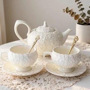 May include: A white ceramic tea set featuring a teapot, two teacups on matching saucers, and two gold-toned spoons. The teapot, cups, and saucers have a textured, embossed surface pattern resembling overlapping petals or leaves.