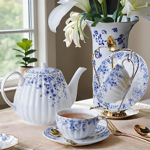 Puede incluir: Un juego de té de porcelana blanca con un patrón floral azul y bordes dorados. El conjunto incluye una tetera acanalada, una taza sobre un plato con una cuchara dorada y una taza a juego en un soporte de metal dorado junto a un jarrón.