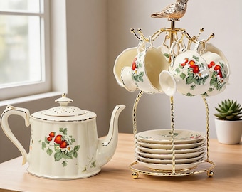 May include: A cream-colored ceramic teapot and a matching set of cups and saucers on a gold-toned metal stand. The teapot and cups feature a strawberry and green leaf pattern with gold trim. A small gold-toned bird figure sits atop the stand.