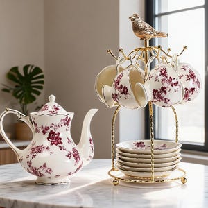 Puede incluir: Una tetera de porcelana blanca y tazas a juego con un estampado floral rojo oscuro. Las tazas cuelgan de un soporte de metal dorado rematado con una figura de pájaro, que también sostiene una pila de platillos a juego en la base.