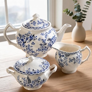 Può includere: Un set da tè in ceramica bianca con un motivo floreale blu. Il set comprende una teiera con coperchio, una zuccheriera con coperchio e due manici, e una lattiera. Ogni pezzo presenta un bordo smerlato e un design botanico blu coordinato.