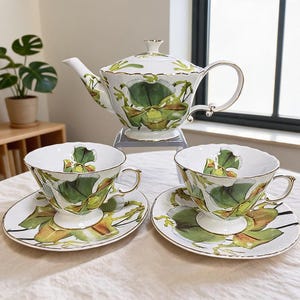 Könnte beinhalten: Ein weißes Porzellan-Teeservice bestehend aus einer Teekanne und zwei Teetassen mit passenden Untertassen. Jedes Teil ist mit einem detaillierten grün-braunen Orchideenmuster verziert und mit einem feinen Goldrand an Kanten und Henkeln versehen.