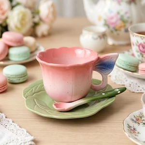 May include: A pink ceramic teacup with a scalloped rim and a blue butterfly wing handle, resting on a green leaf-shaped saucer. A matching green ceramic spoon with white dots rests inside the cup, completing this floral-inspired tea set.