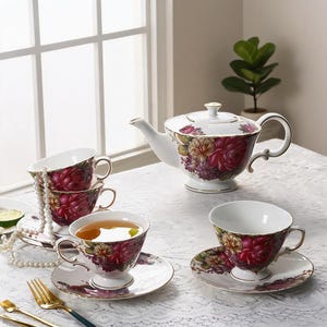 Puede incluir: Un juego de té de porcelana blanca con un vibrante estampado floral de rosas rojas oscuras y amarillas. El conjunto incluye una tetera con tapa, tres tazas con sus platos a juego y un collar de perlas blancas sobre las tazas apiladas.