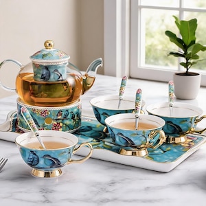 Puede incluir: Un juego de té que incluye una tetera de cristal con estampado de pájaros y flores, un calentador a juego, tres tazas con asas doradas y tres cucharas a juego. El conjunto está dispuesto sobre una bandeja rectangular con motivos florales.