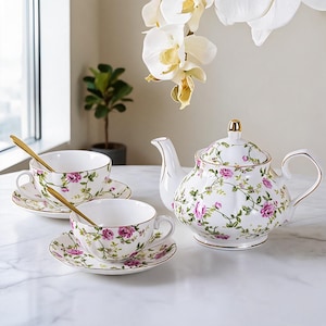 Puede incluir: Un juego de té de porcelana blanca con un delicado patrón de rosas rosadas y enredaderas verdes con borde dorado. El conjunto incluye una tetera con tapa, dos tazas con sus platos a juego y dos cucharas doradas sobre una superficie de mármol.