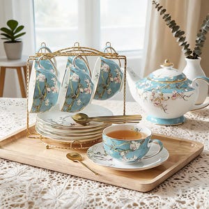 Puede incluir: Un juego de té que incluye una tetera, tazas y platillos de porcelana azul claro decorados con patrones de flores de almendro blancas y rosas. El set incluye un soporte de metal dorado para tazas y platillos, junto con pequeñas cucharas doradas.