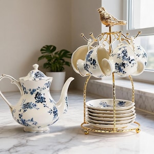 Puede incluir: Una tetera de porcelana blanca y un juego a juego de tazas y platillos decorados con motivos florales azules. Las tazas cuelgan de un soporte de metal dorado rematado con una figura de pájaro, mientras los platillos se apilan en la base.