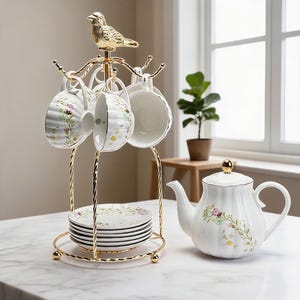 May include: A gold-toned metal stand topped with a bird figurine, holding white ceramic teacups with floral patterns. A stack of matching floral saucers sits at the base of the stand, accompanied by a white ceramic teapot with a gold lid knob.