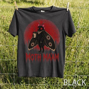 Può includere: Una t-shirt nera con la grafica di una falena dagli occhi rossi, un fiocco rosa e ali con motivi circolari. La falena tiene in mano una tazza di caffè su uno sfondo circolare rosso. Il testo MOTH MA'AM è stampato in rosso sotto la grafica.