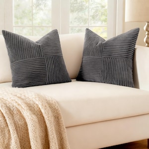 May include: Two gray corduroy throw pillows with a patchwork design rest on a cream-colored sofa. The pillows have a textured, ribbed appearance. A beige knitted throw blanket is draped over the sofa.