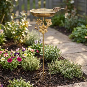 May include: An antique-style bird bath with a shallow, circular basin. The bird bath is made of gold-coloured metal and features floral and butterfly embellishments. A small bird is perched on the edge of the basin. The bird bath is set in a garden.