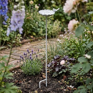 Könnte beinhalten: Ein weißes Vogelbad aus Metall mit einem kleinen Vogel auf dem Rand, auf einem hohen Pfahl. Das Vogelbad befindet sich in einem Garten mit Blumen und Pflanzen. Der Pfahl hat eine zweizinkige Basis zum Einsetzen in den Boden.