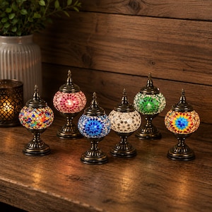 May include: Six small table lamps with rounded mosaic glass shades in various colors including red, blue, green, and white. Each lamp features a dark bronze-toned metal base and a pointed finial top, arranged in a group on a wooden surface.