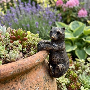 Op de afbeelding: Een bronzen beerfiguur die zich vastklampt aan de rand van een terracotta bloempot. De beer heeft getextureerde vacht en is omgeven door groene vetplanten, met een vage achtergrond van lavendel en roze bloemen.