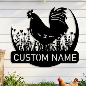 Könnte beinhalten: Schwarzes Metall-Hahnschild mit dem Text "CUSTOM NAME" auf weißem Holzgrund. Das Schild zeigt eine Hahn-Silhouette, umgeben von Gras und Blumen. Eine Henne und Küken sind im Vordergrund.