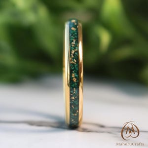 Pode incluir: Um anel de ouro com uma faixa central de malaquite verde e flocos de ouro. O anel está sobre um fundo verde desfocado e uma superfície de mármore branco. Close-up do anel.