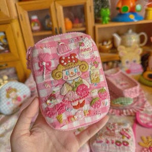 May include: A rectangular pink and white gingham zipped pouch featuring embroidered designs of a central character with blonde hair and a red bow, surrounded by various sweets, desserts, and strawberry motifs on the front surface.