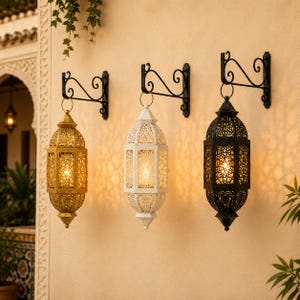 Puede incluir: Tres faroles de pared en acabados dorado, blanco y negro, cada uno con intrincados patrones de metal recortado y una luz interna cálida. Los faroles cuelgan de soportes de metal negro con detalles de volutas en una pared clara.