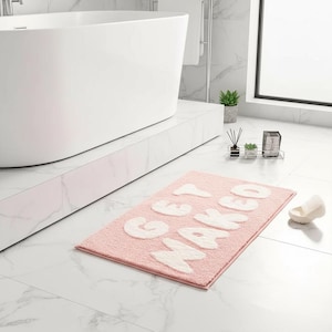 May include: A rectangular light pink bath mat with a textured surface featuring the white text GET NAKED in a bold, rounded font. The mat is placed on a white marble floor in front of a white bathtub in a bathroom setting.