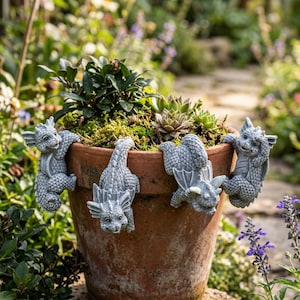 Puede incluir: Una maceta de terracota con cuatro figuras de dragones grises aferradas al borde. La maceta está llena de vegetación, incluyendo suculentas y musgo. El fondo muestra un camino de jardín y flores.