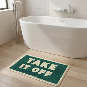 Puede incluir: Una alfombra de baño rectangular verde con las palabras "TAKE IT OFF" en letras blancas. La alfombra tiene un borde color crema y está colocada sobre un suelo de madera clara frente a una bañera blanca.